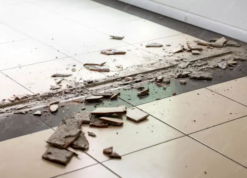 Broken and chipped floor tiles scattered across a tiled bathroom floor, showing damaged surface underneath.