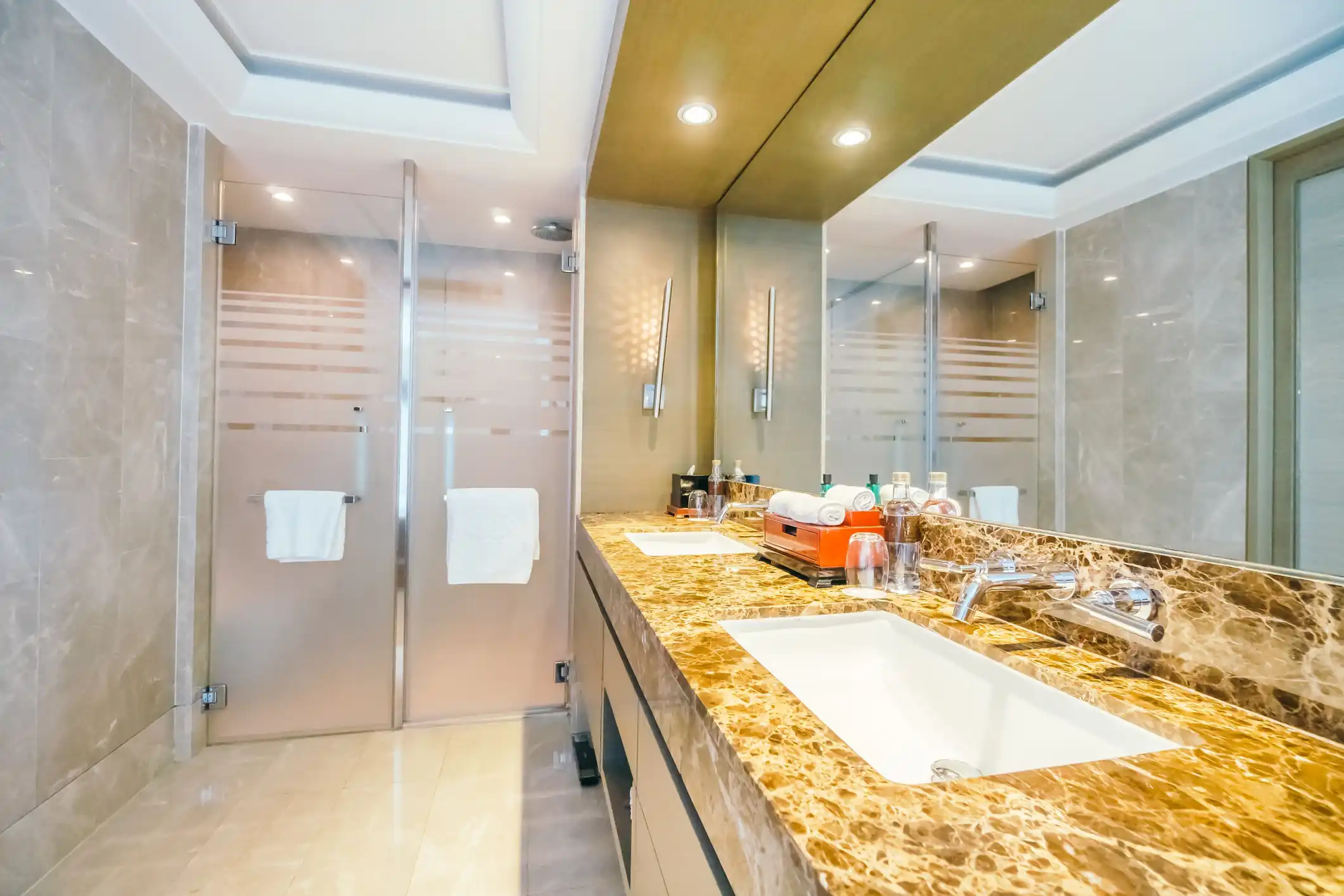A well-lit bathroom with marble floor and wall tiles, double vanity sinks, and a glass shower enclosure.